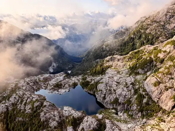 An aerial view of a dark alpine lake in pale rocky ground, with mist drifting through the valley