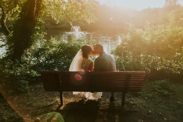A bride and groom kissing on a park bench in golden evening light between trees