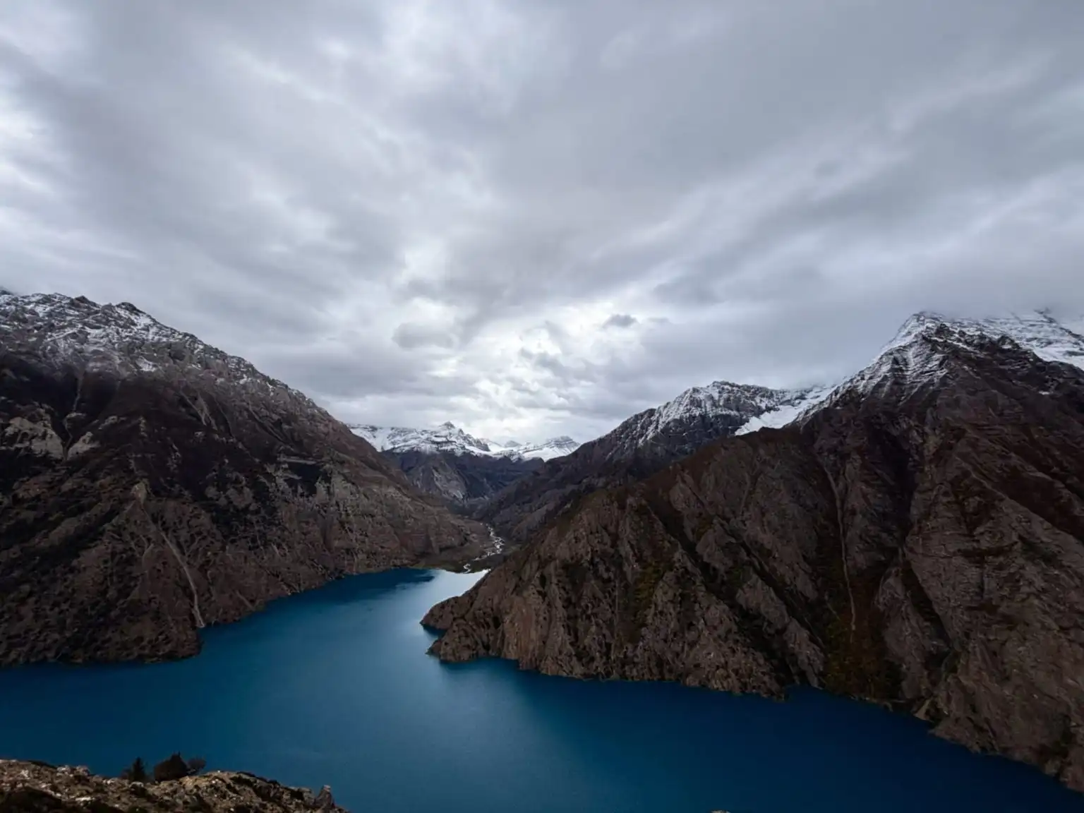 A deep blue lake winding between steep mountains with snow on the peaks, under grey cloud
