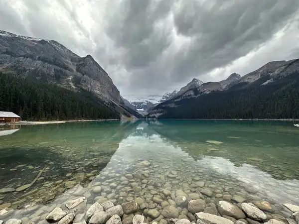 Clear lake water over smooth stones, reflecting forested mountains under storm clouds