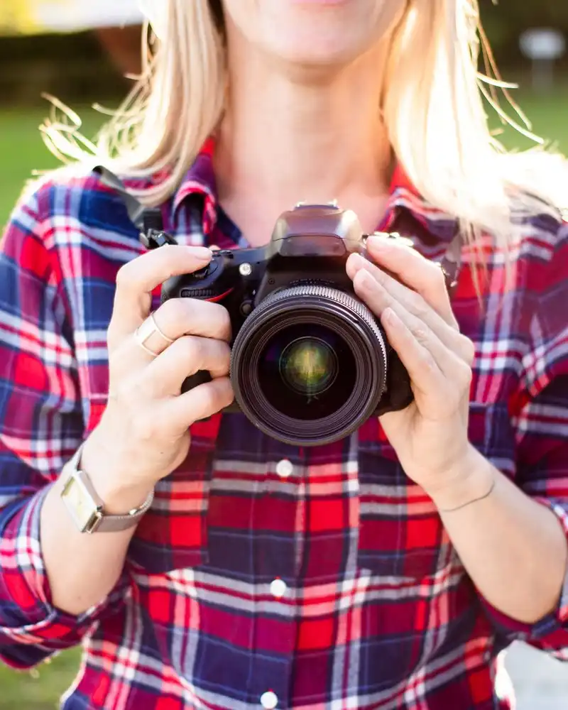 A woman in a plaid shirt holding a camera with both hands, outdoors in afternoon light
