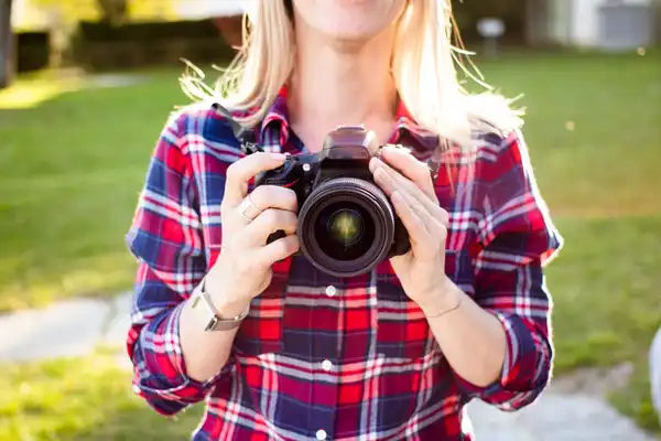 A woman in a plaid shirt holding a camera with both hands, outdoors in afternoon light