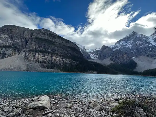 A turquoise mountain lake below snow-dusted rock peaks, with a stony shore in front
