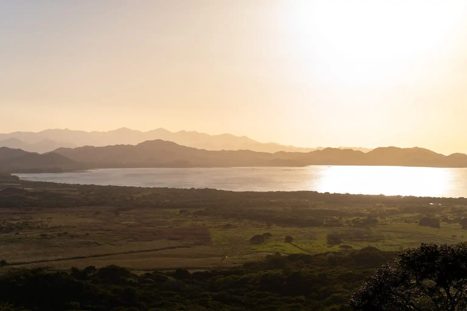 Low sun over a wide lake and layered hills, with open fields in the foreground