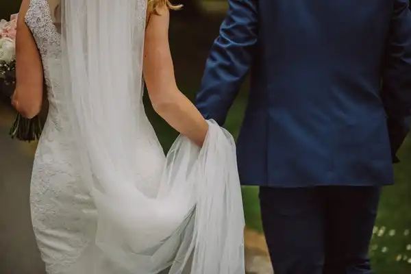 A bride in a lace dress and long veil walking arm in arm with a groom in a navy suit